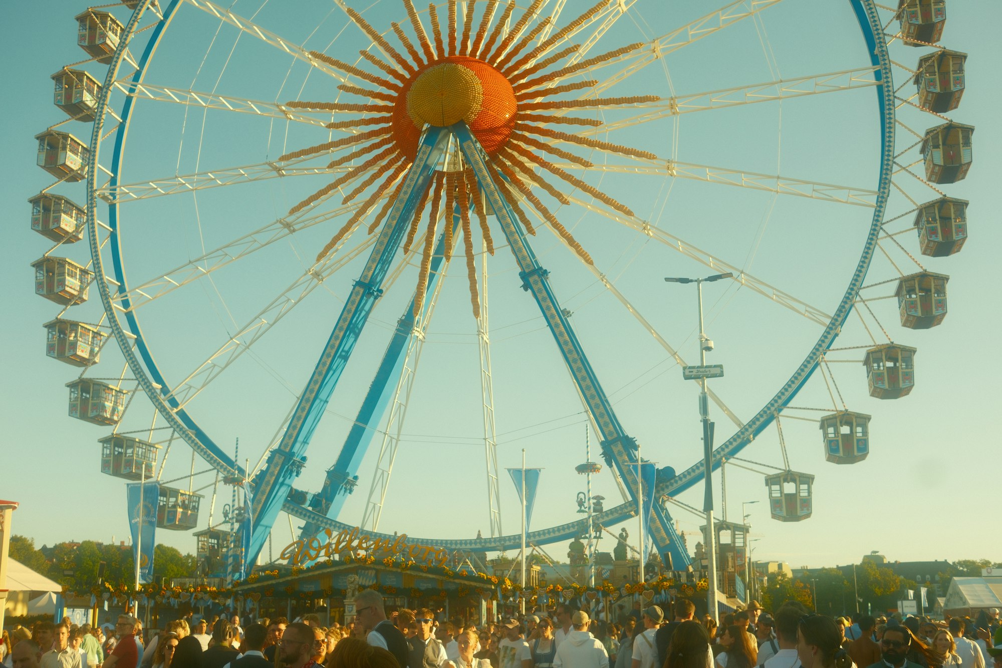 A ferris wheel and crowd gathered at an outdoor community festival on a sunny day.